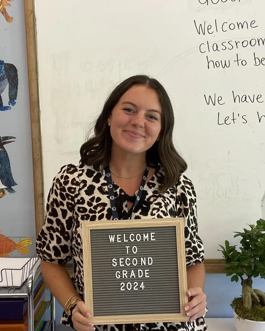 Maya smiling in her classroom, holding a Welcome to Second Grade letterboard